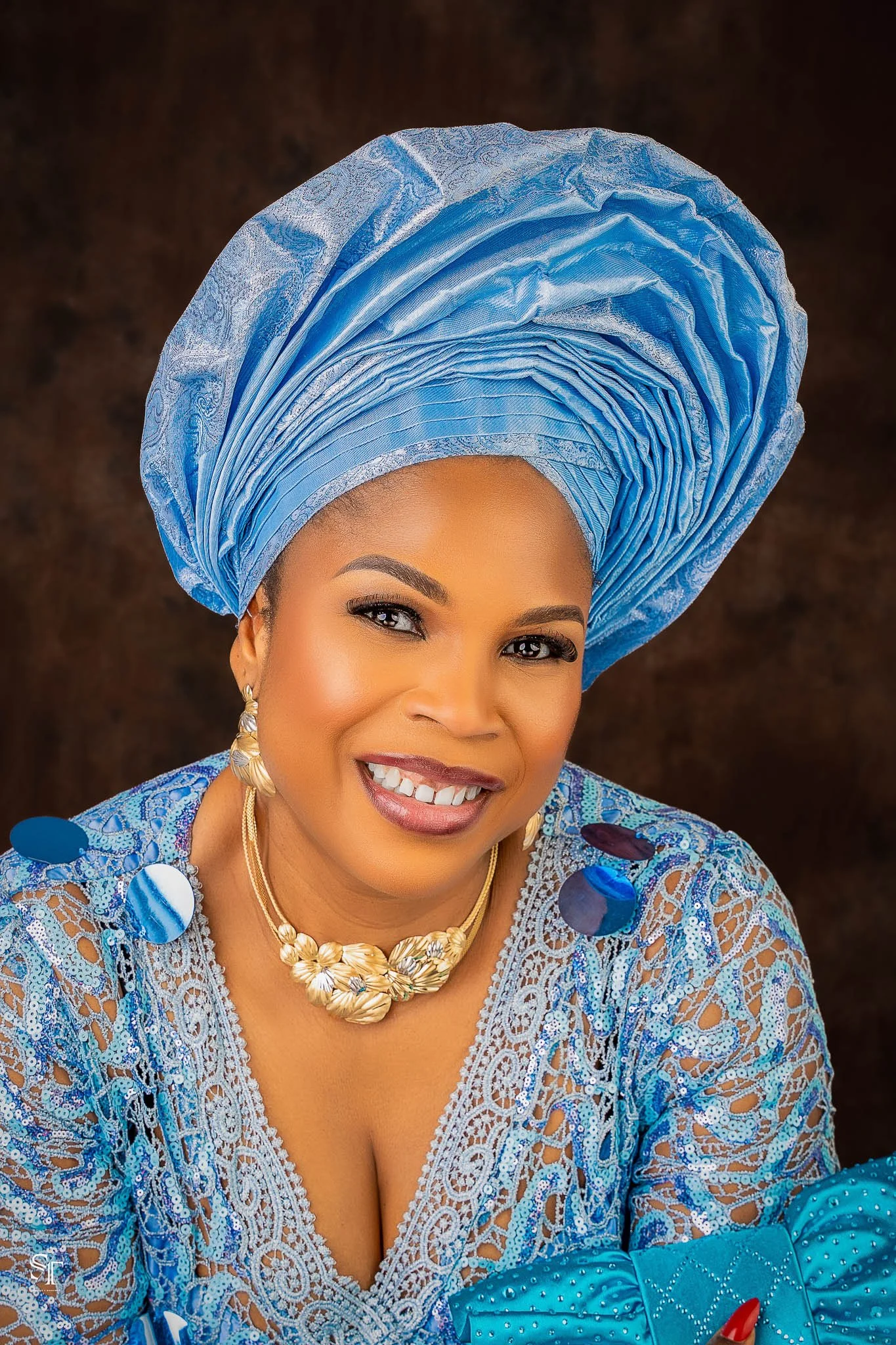 A woman dressed in traditional Nigerian attire, wearing an elaborate blue gele (headwrap) and a matching lace outfit, with gold jewelry and a confident smile.