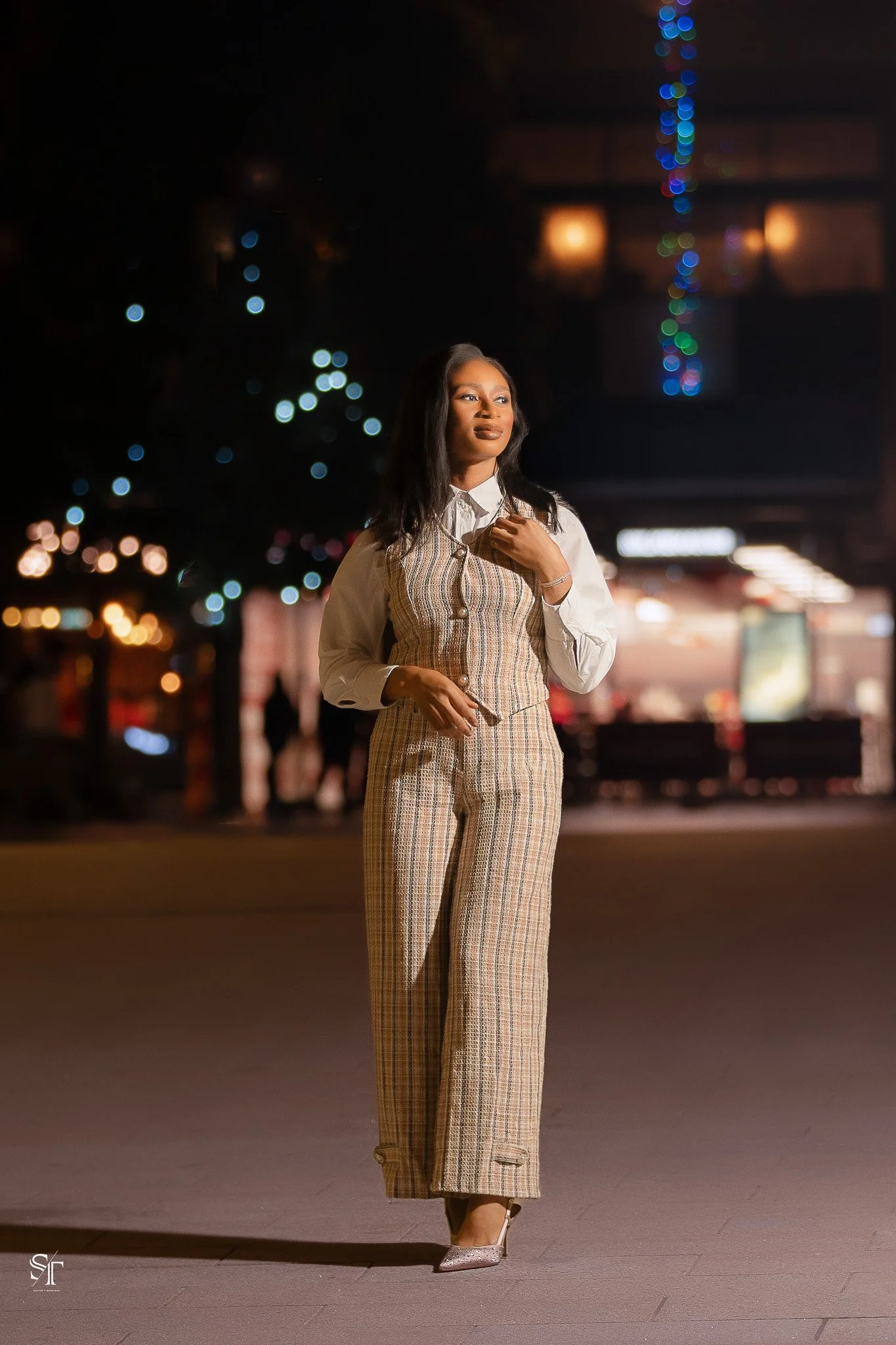 A woman in stylish plaid pants and a vest, standing outdoors at night with blurred city lights and string lights in the background.