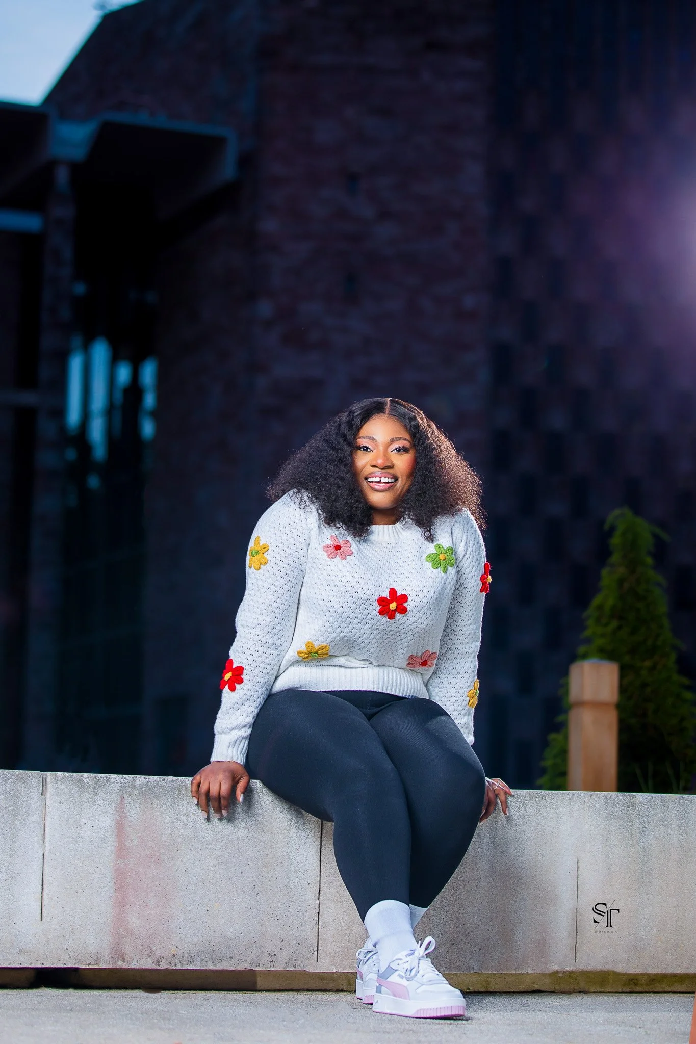 A woman sitting on a concrete ledge outdoors during evening, wearing a gray sweater with colorful flower appliques, black leggings, white sneakers, and smiling at the camera.