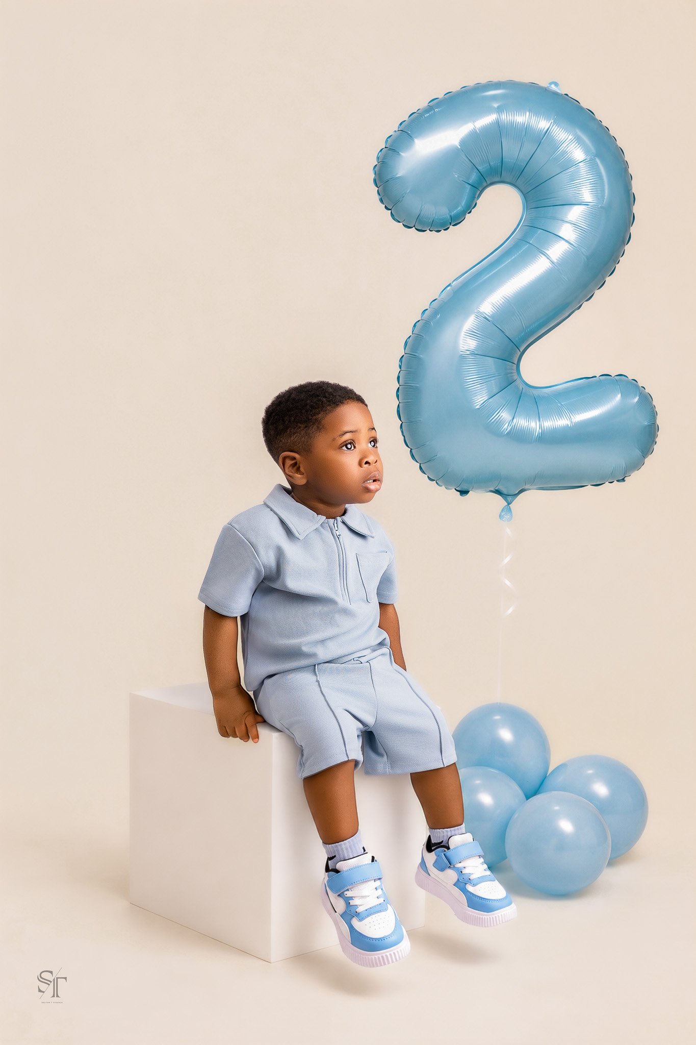 A young boy sitting on a white cube, wearing a light blue outfit and sneakers, with a large blue number 2 balloon behind him, and several smaller blue balloons on the floor.