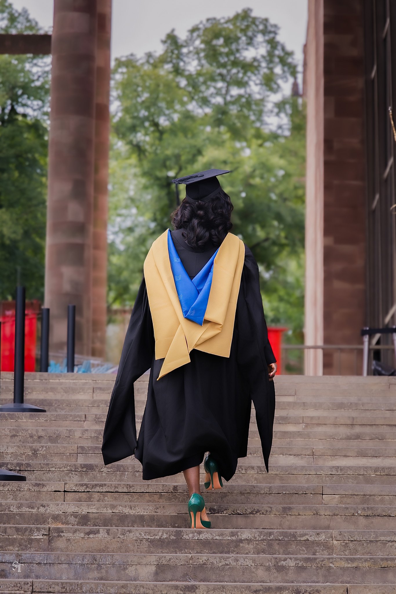A woman in graduation cap and gown with a yellow and blue stole, walking up the steps outside a building, seen from the back. She wears green heels.