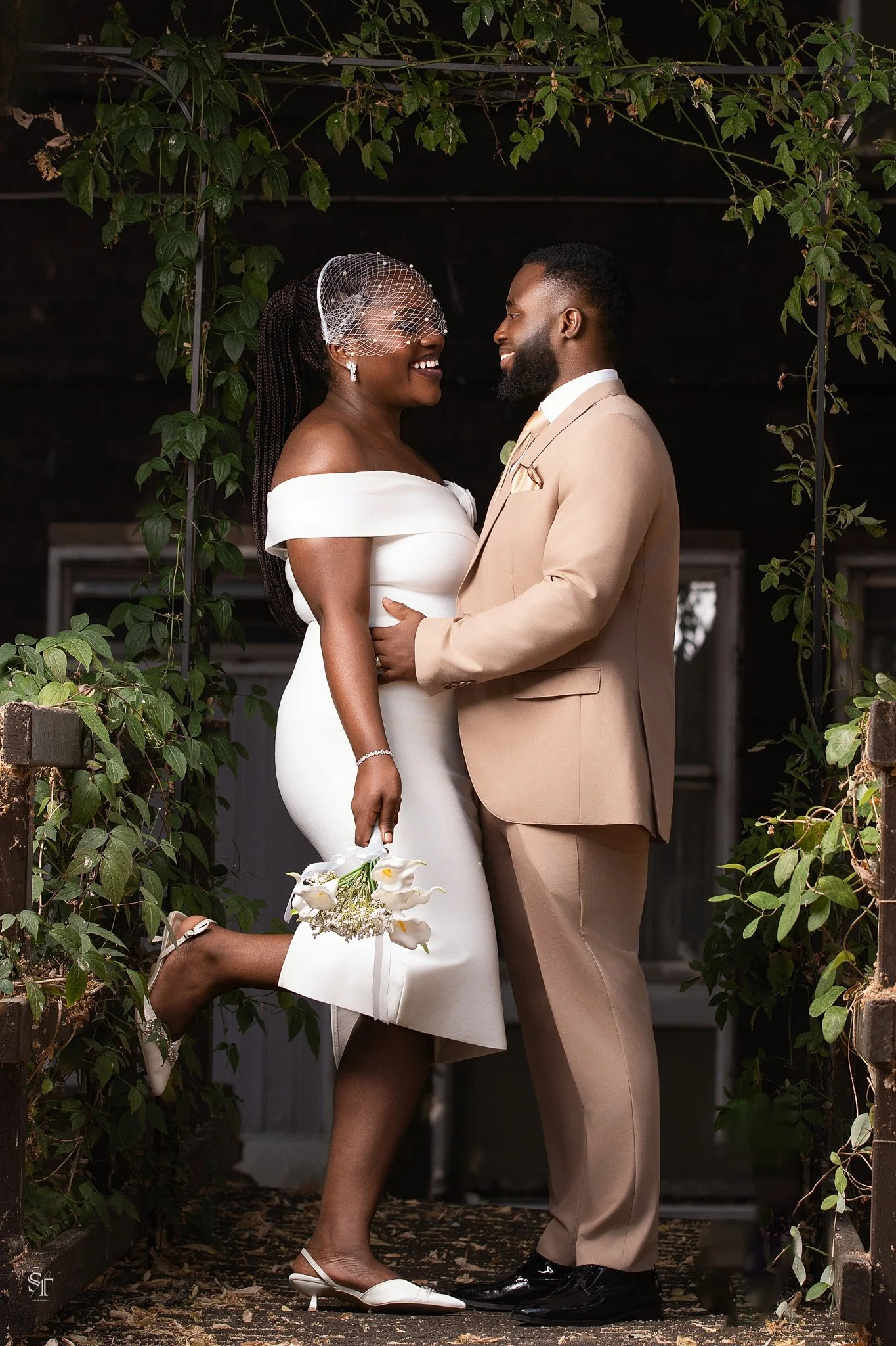 A couple dressed in wedding attire gazing into each other's eyes, standing in a garden archway covered with green leaves.
