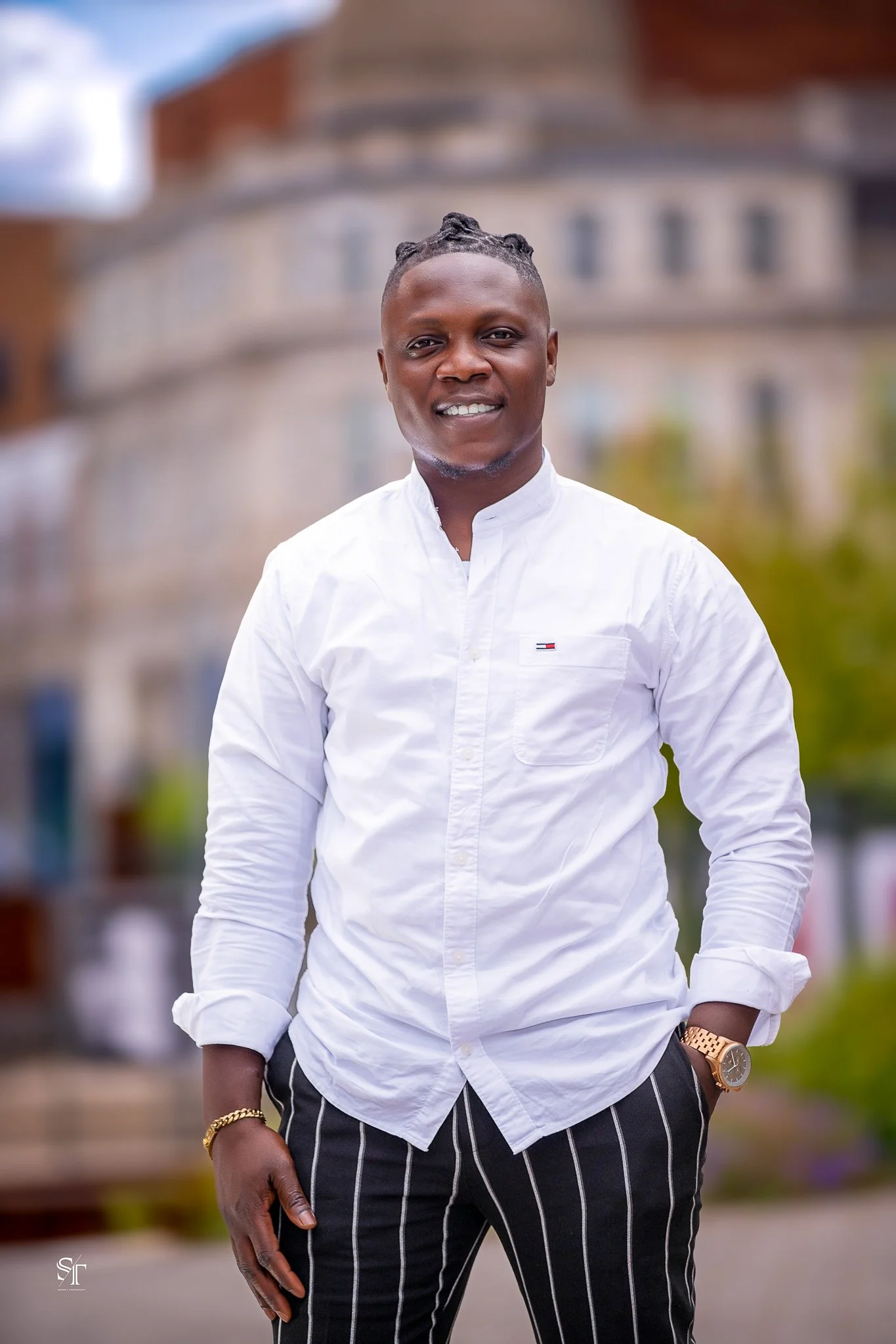 A young man with dark skin and short patterned hair, smiling and standing outdoors. He is wearing a white button-up shirt, black and white striped pants, a gold watch on his left wrist, and a gold bracelet on his right wrist. The background features 