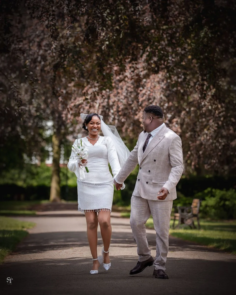 A joyful bride and groom walking hand in hand outdoors, surrounded by trees and greenery, with the bride holding a bouquet of white flowers.