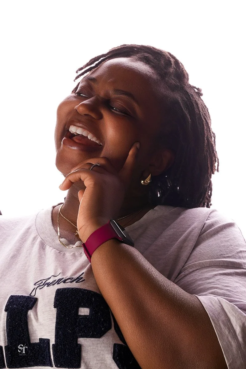 A woman with short dreadlocks, smiling with her eyes closed, holding her face with one hand, wearing a watch, earrings, and necklaces, with a white t-shirt and a white background.