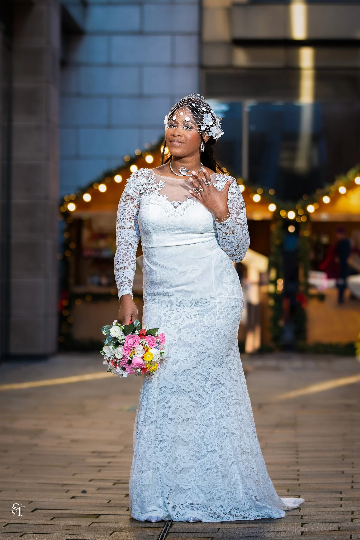 A bride in a white lace wedding gown holding a bouquet of pink and white flowers, standing outdoors with festive holiday decorations in the background.