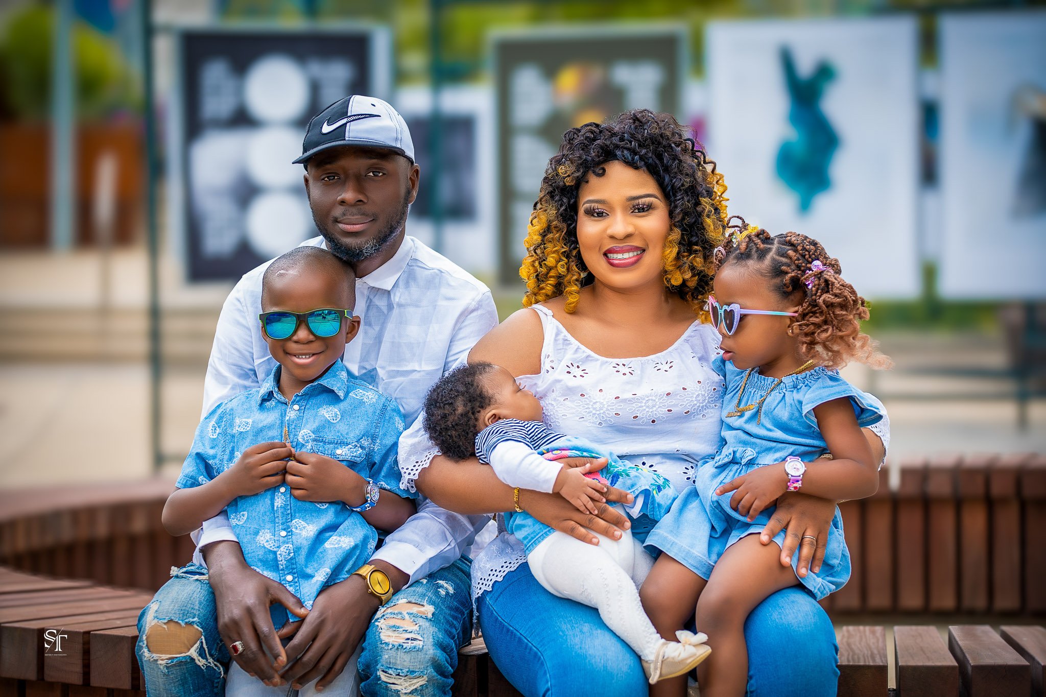 Family of five sitting on a park bench, smiling, with children wearing blue clothes and sunglasses, the mother holding a baby, father and two children around her.
