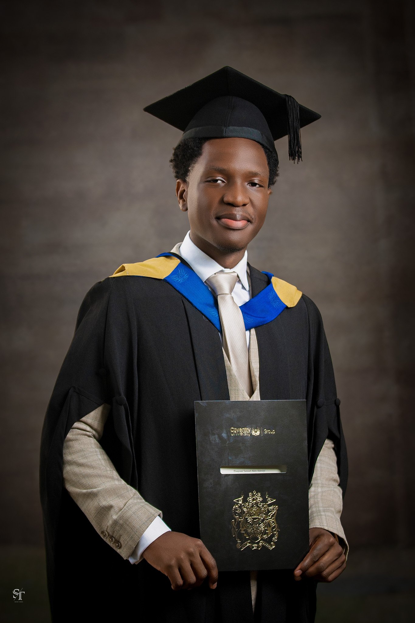 A young man in a graduation cap and gown holding a diploma with a gold emblem, standing against a dark background.