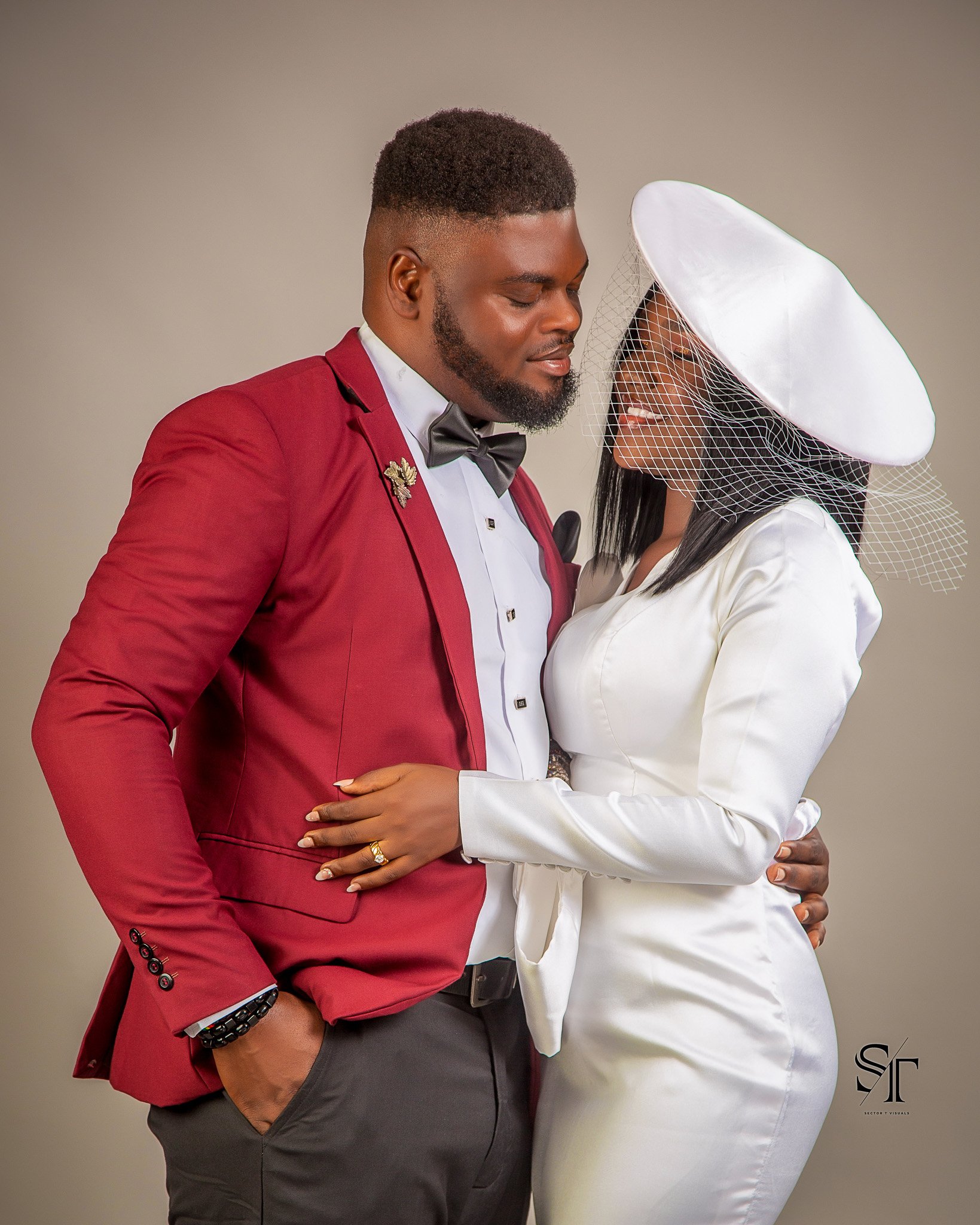 A couple dressed in formal wedding attire, embracing and smiling at each other, against a plain background.