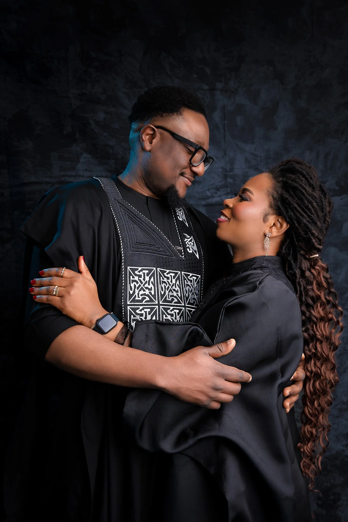 A couple dressed in traditional African attire, embracing and looking into each other's eyes against a dark backdrop.