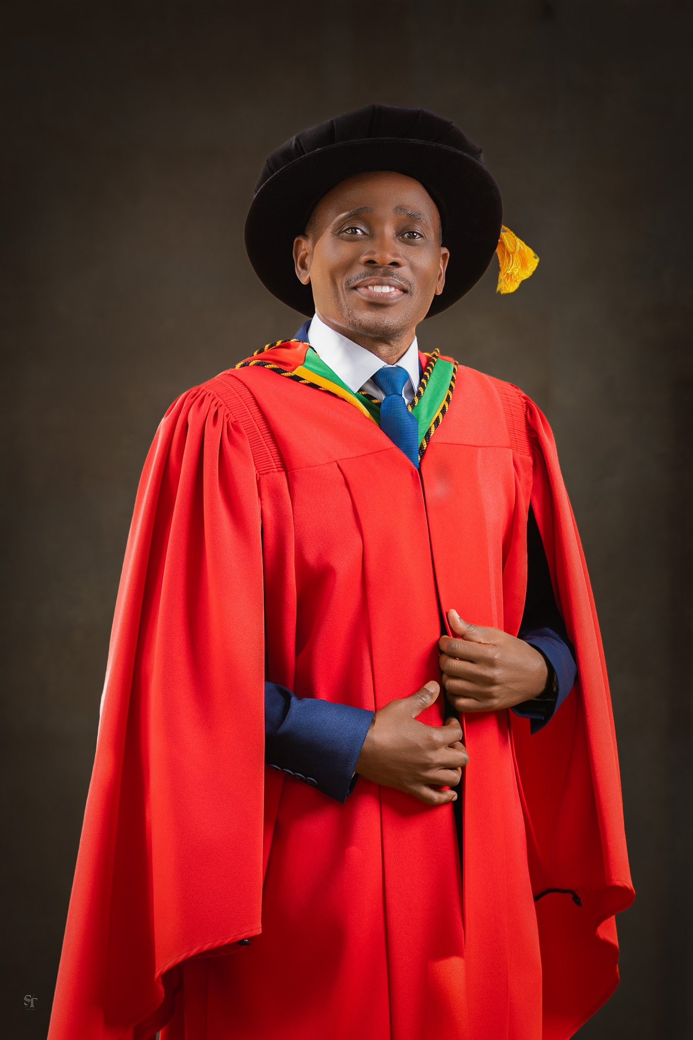 Man wearing academic regalia, black hat with yellow tassel, red gown, and a green, yellow, black, and white hood, posing against a dark background.