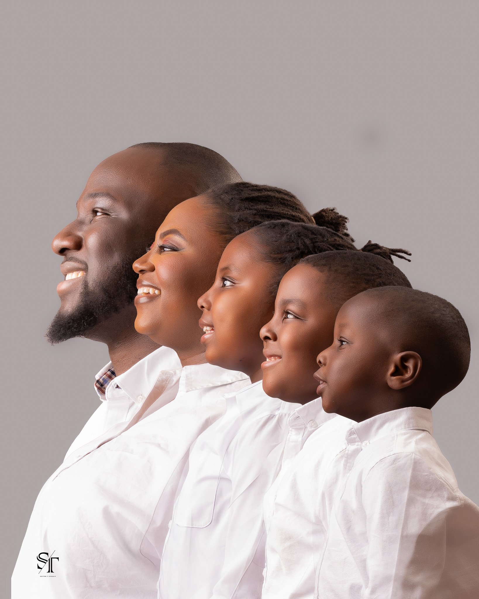 A group of five diverse people standing in a family line, smiling and looking to the left, against a gray background, wearing white shirts.