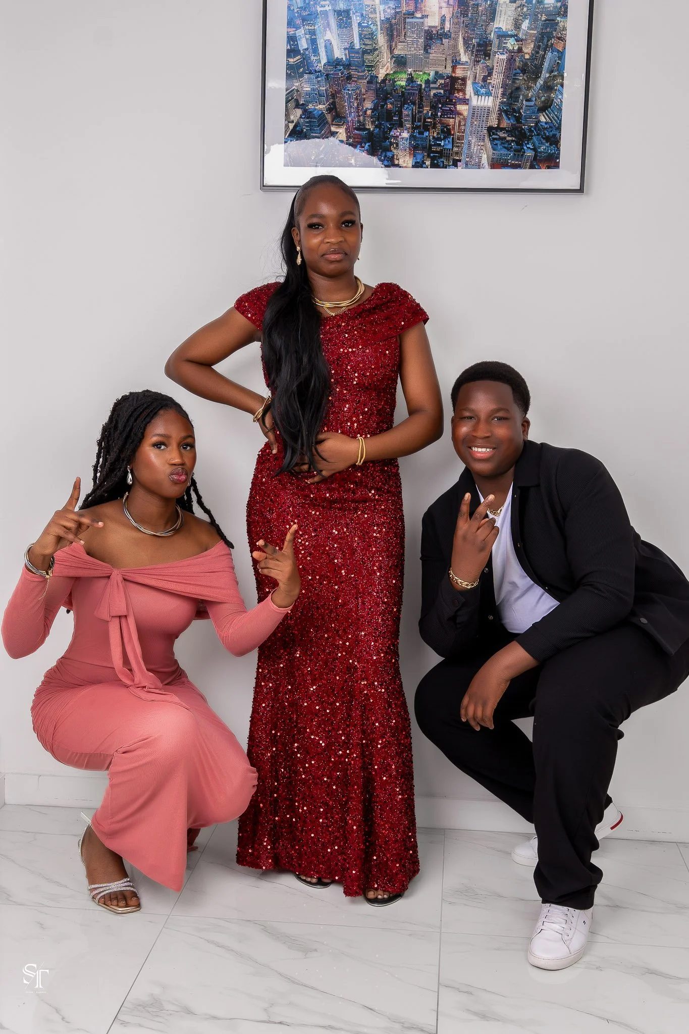 Three young adults, two women and one man, posing in a modern indoor setting with a white wall and a cityscape framed picture behind them. The woman in the middle wears a red, floor-length, sequined dress. The woman on the left squats in a pink dress