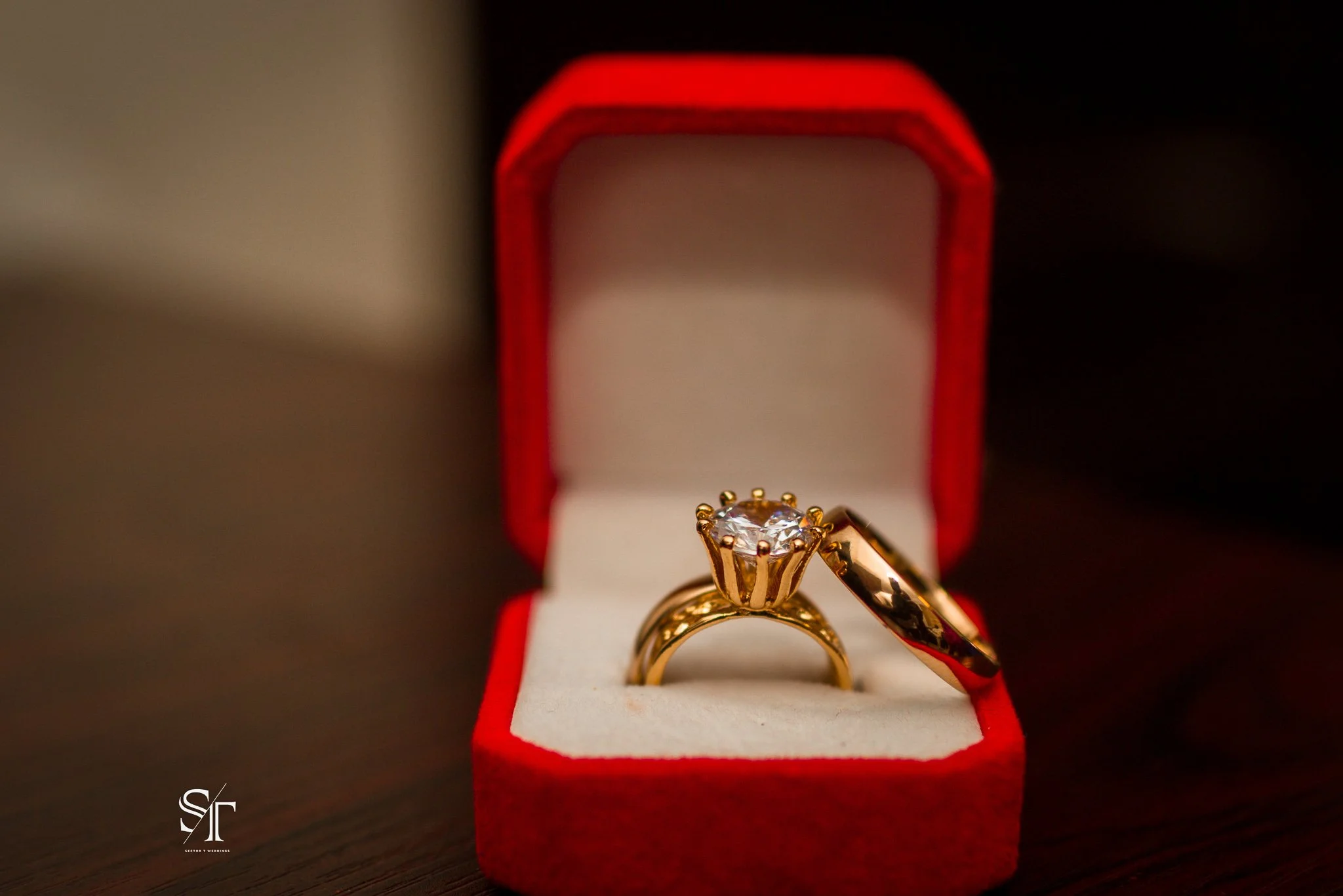 A gold engagement ring with a large central diamond in a red velvet box lined with white fabric.