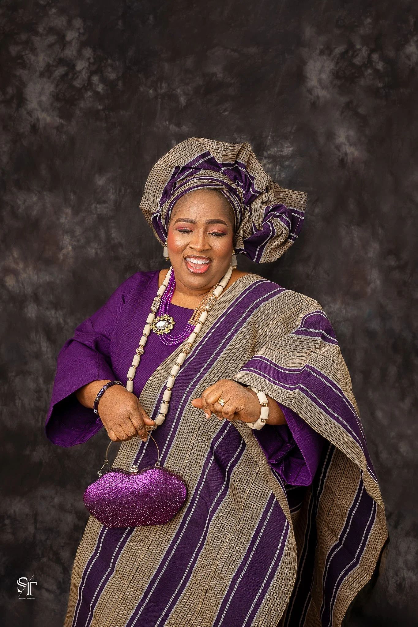 A woman dressed in traditional Nigerian attire, smiling and posing with a purple heart-shaped purse, wearing a purple and beige striped headwrap and matching dress, against a dark textured background.