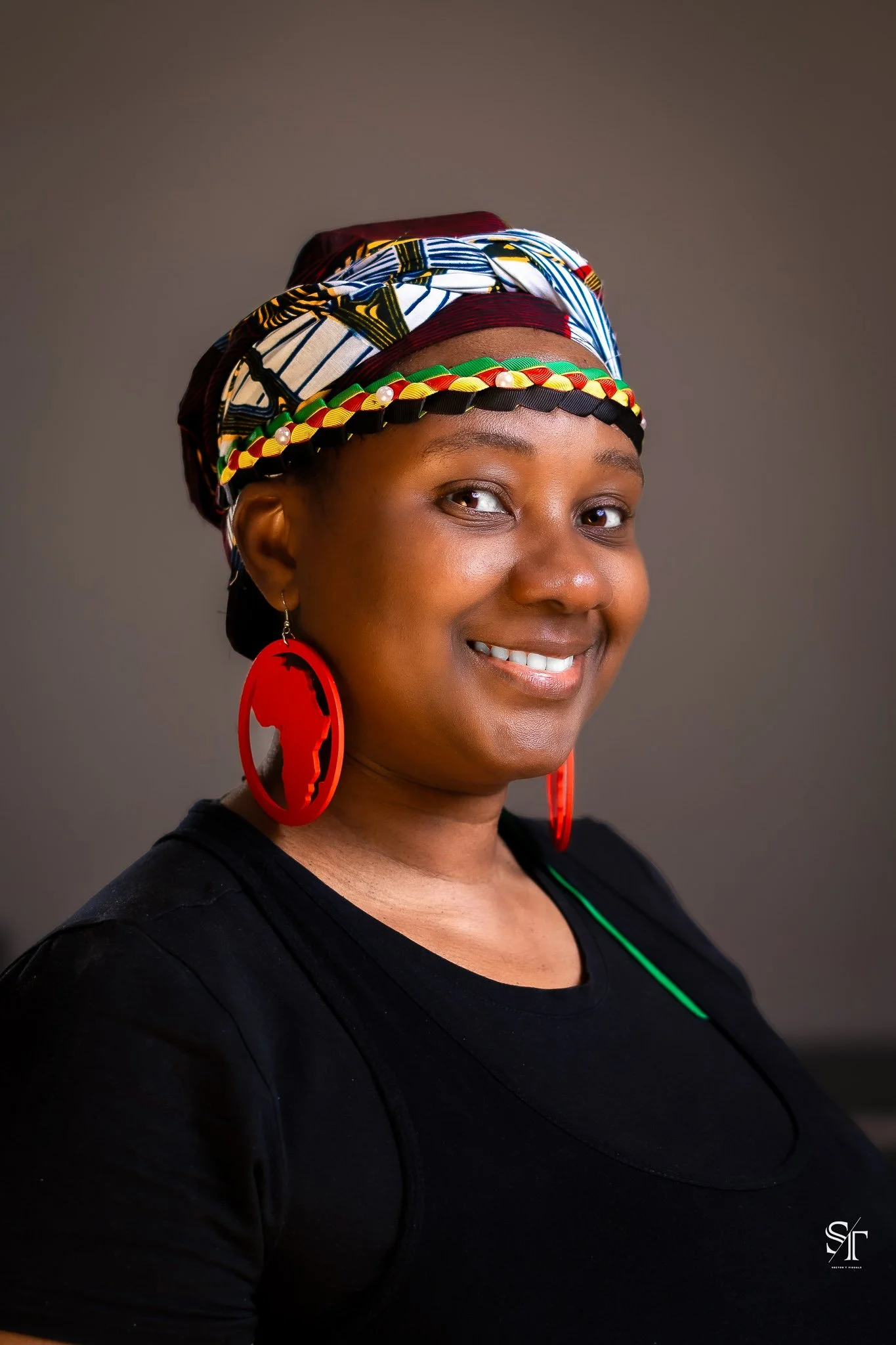 A smiling woman with dark skin, wearing large red earrings featuring a silhouette of the African continent, a colorful patterned headwrap, and a black top, against a plain, gradient background.