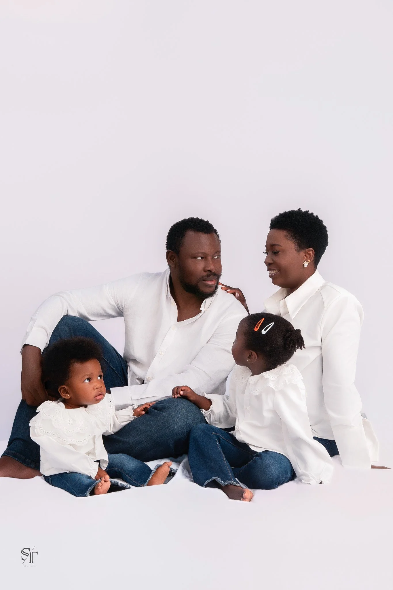 An African American family of six sitting on a white background, wearing white shirts and blue jeans. The father and mother are seated with three children, two girls and one boy, engaging in conversation and affection.