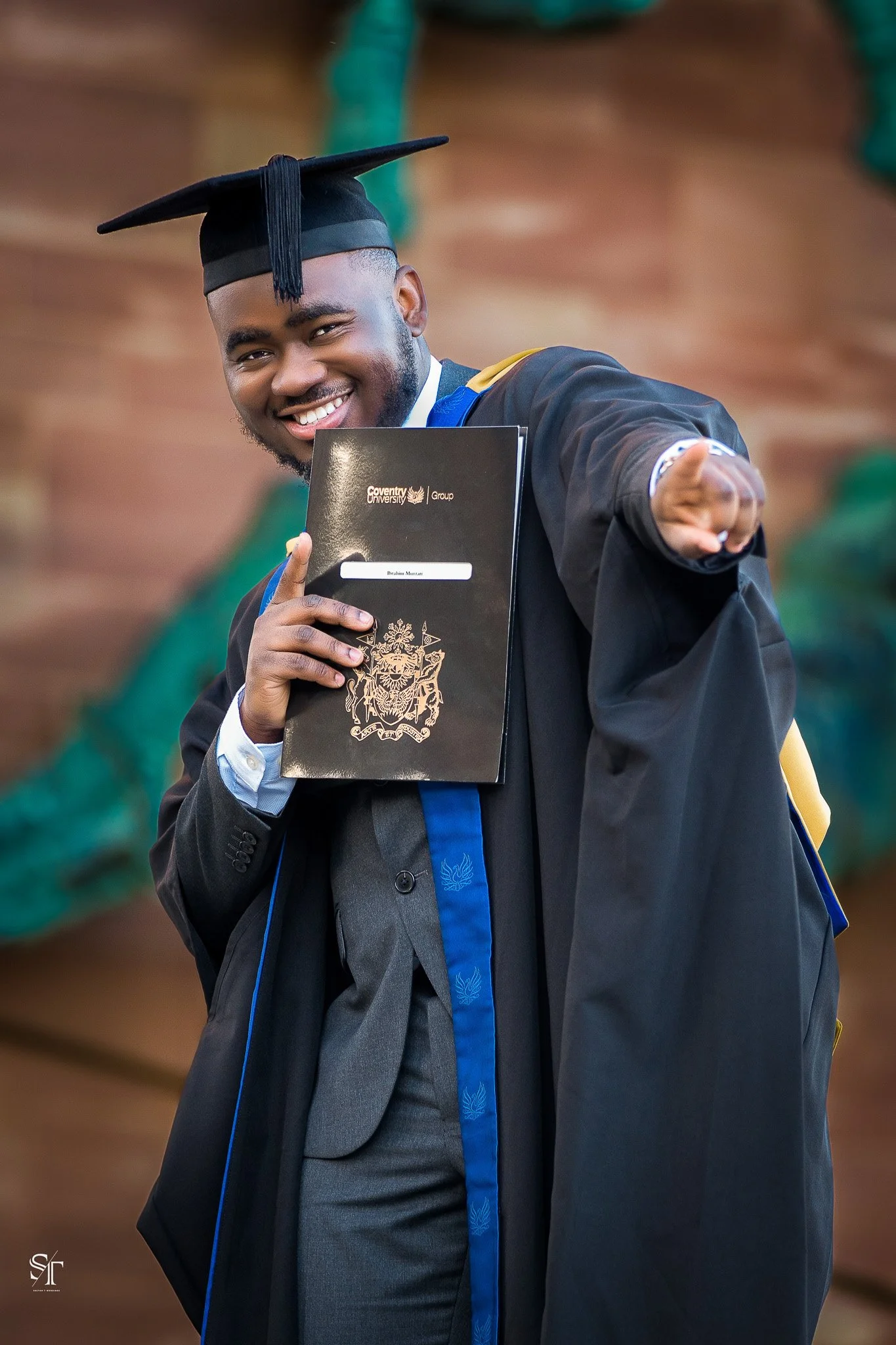 A happy young man dressed in graduation cap and gown, holding a diploma and pointing towards the camera.