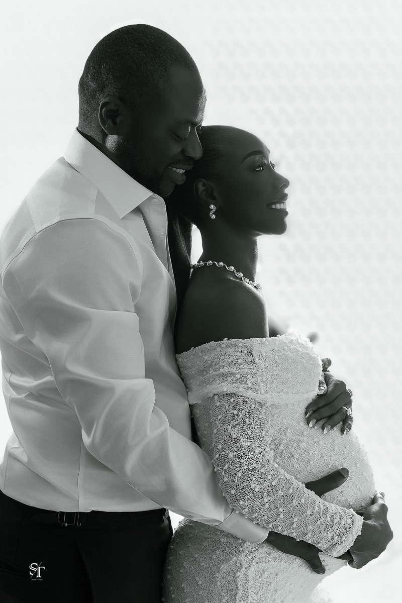 A black and white photo of a couple, with the man embracing the woman from behind, both smiling, with the woman wearing a lace off-shoulder gown and jewelry, celebrating a special moment.