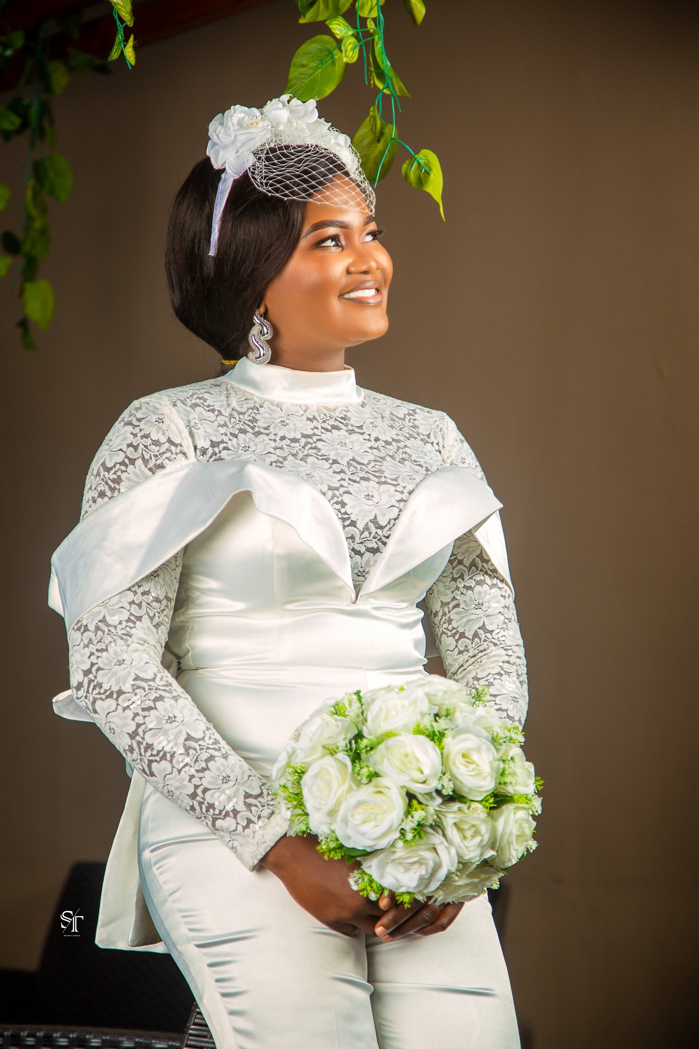 A woman in a white wedding dress, holding a bouquet of white roses, standing under green hanging plants, smiling, wearing lace sleeves and earrings.