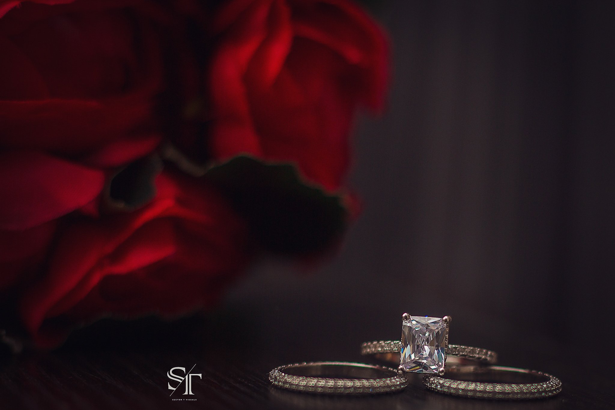 A close-up of a sparkling square-cut diamond engagement ring placed between two thin, diamond-studded wedding bands on a dark surface, with a blurred red flower in the background.