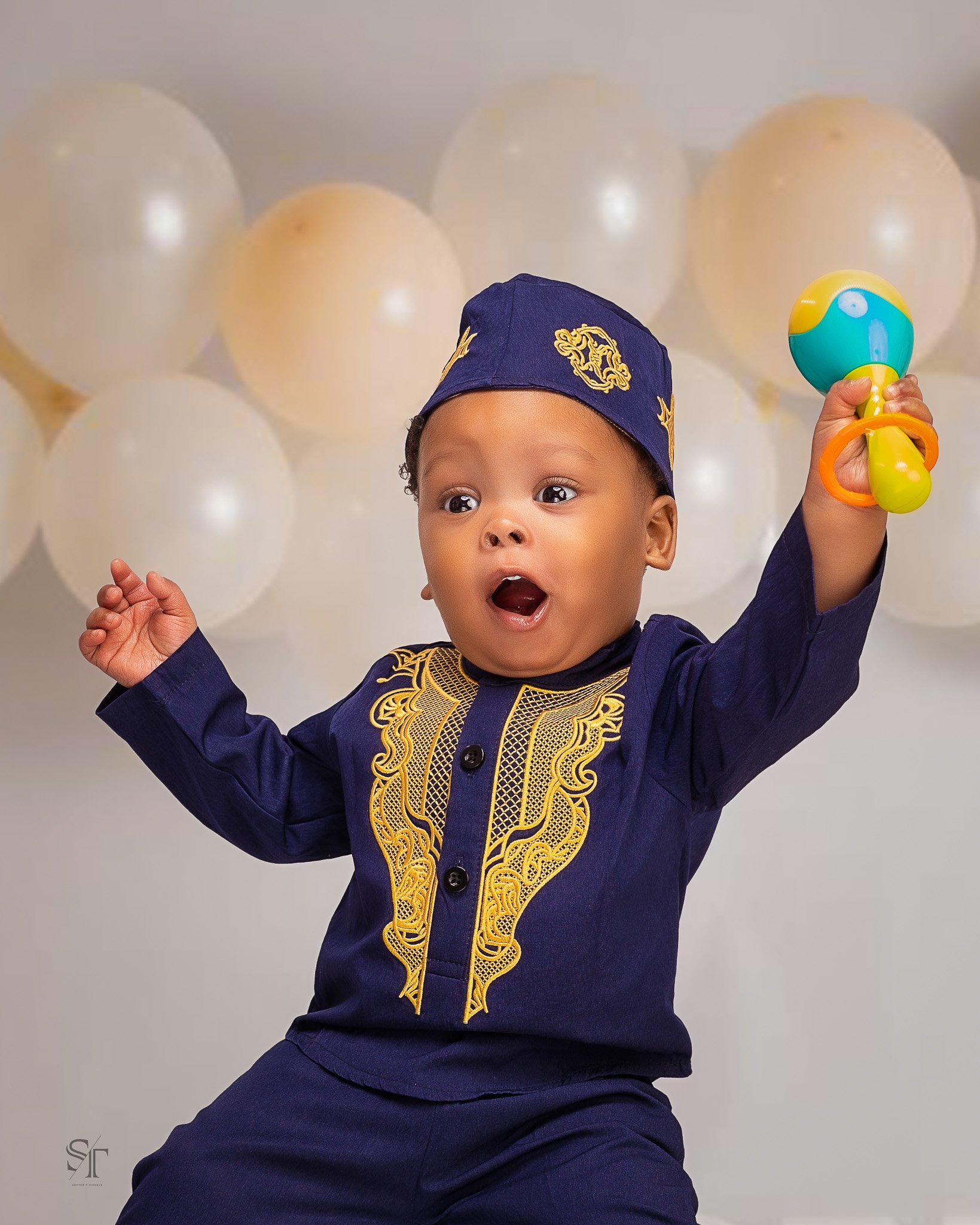 A young child dressed in a navy blue traditional outfit with gold embroidery, wearing a matching navy cap, holding a colorful toy, with white balloons in the background.