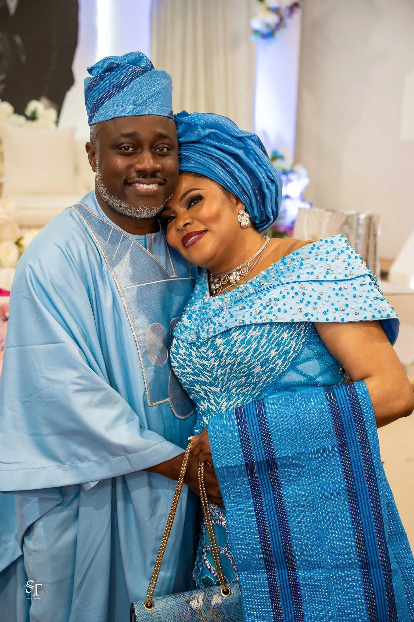 A smiling couple dressed in traditional African attire, embracing at a celebration or wedding, with a decorated background.