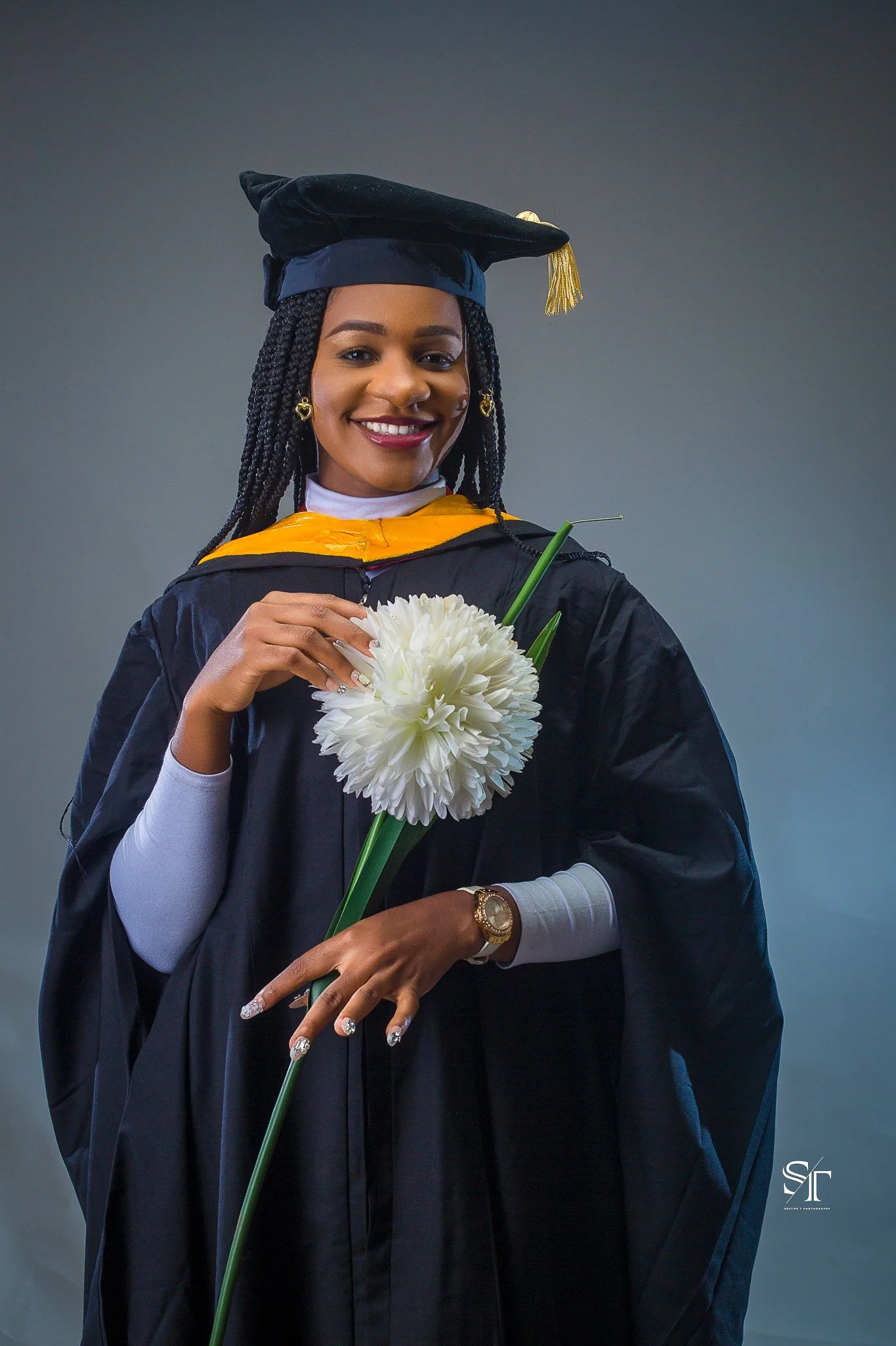 A woman in graduation cap and gown holding a white flower, smiling for a portrait.