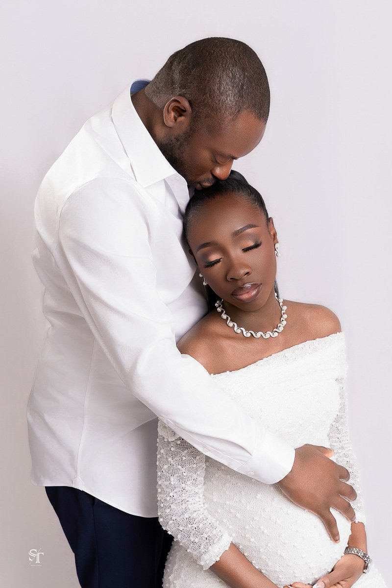 A man and a pregnant woman embracing, with the man gently resting his head on her forehead. Both are dressed in white, with the woman wearing a lacy off-shoulder dress and pearl jewelry, and the man dressed in a white shirt. They appear to be in a st