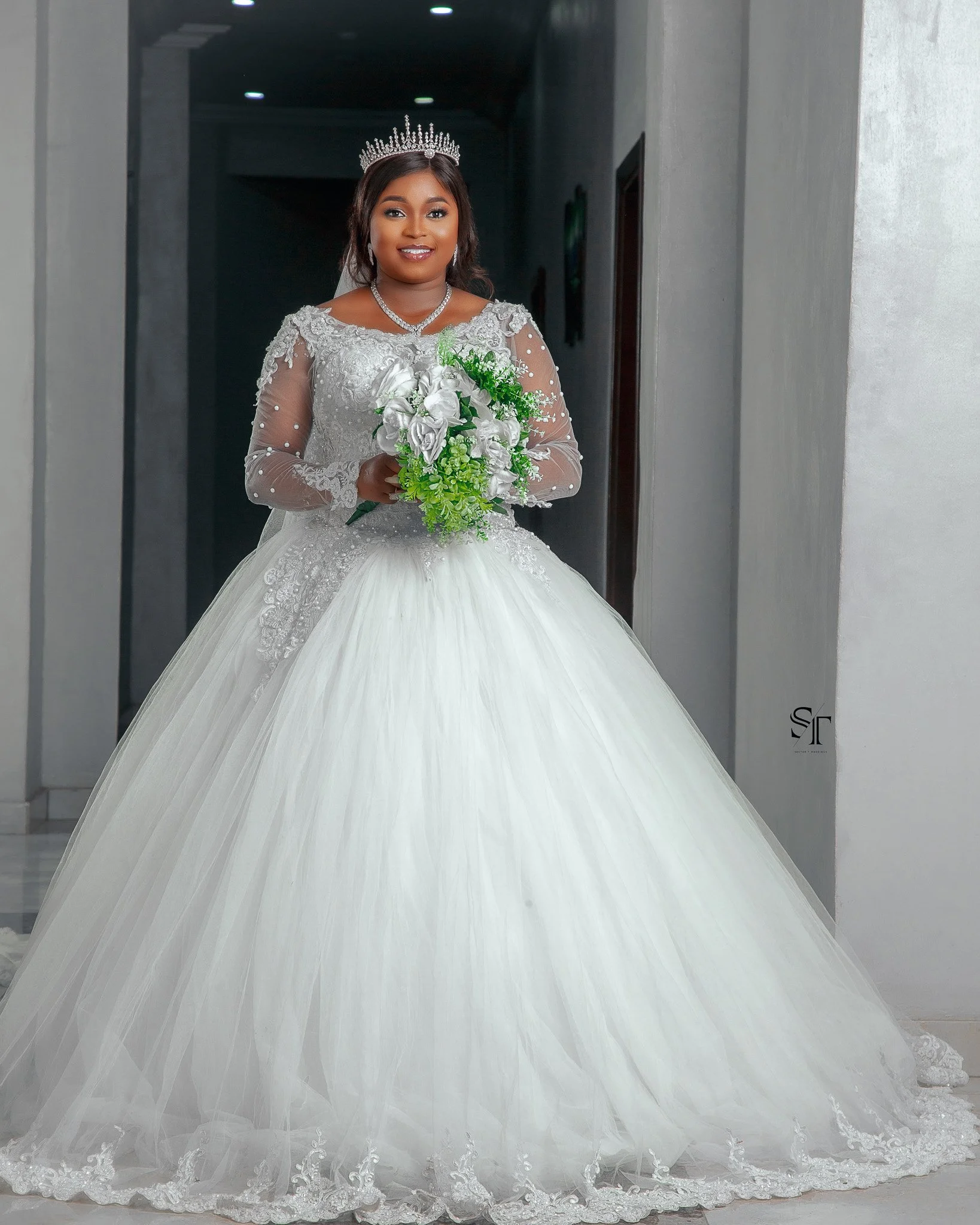 A bride in a white wedding gown holding a bouquet of white and green flowers, wearing a tiara, standing in a hallway.