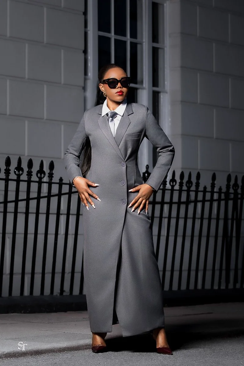 A woman in a tailored grey suit, wearing sunglasses, standing confidently outside at night in front of a black iron fence and a grey building.