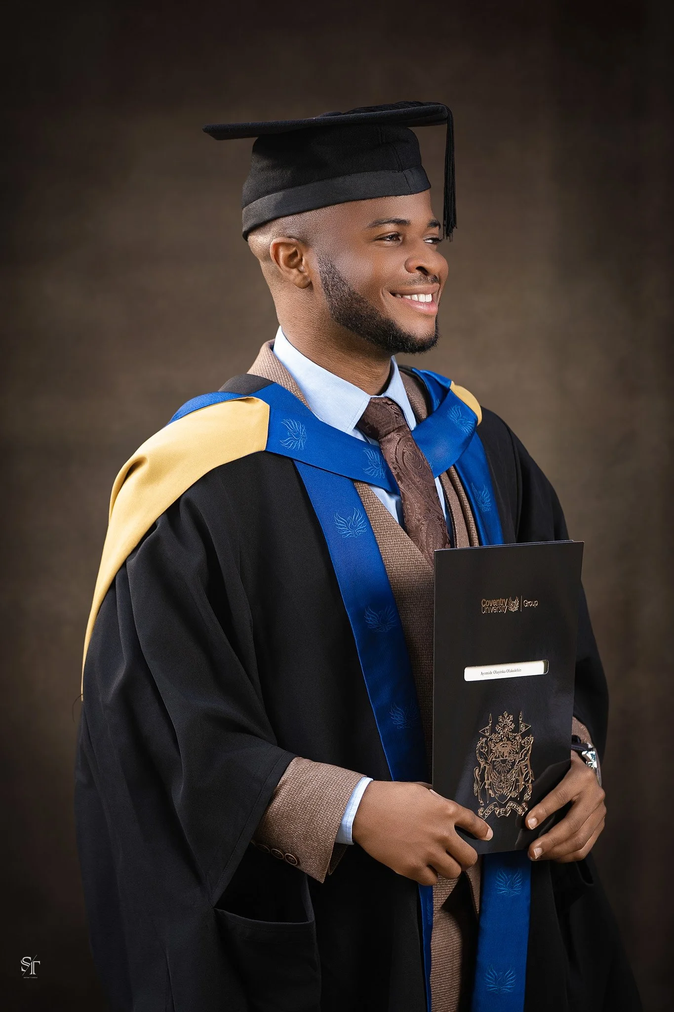 Graduation portrait of a young man in cap and gown holding a diploma, smiling, against a dark background.
