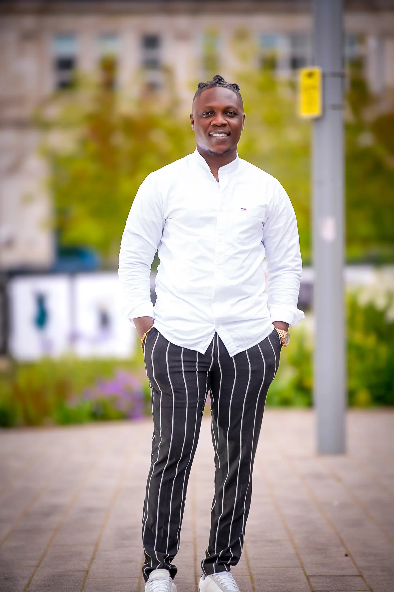 Man wearing a white long sleeve shirt, striped black and white pants, and white sneakers, standing outdoors with hands in pockets, smiling, with a blurred background of trees, buildings, and a street sign.