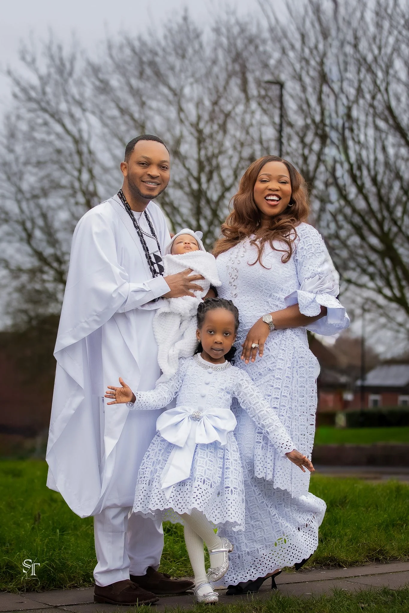 A family of five dressed in white poses outdoors on a cloudy day, with leafless trees in the background. The father holds a newborn baby, while a young girl stands in front, and the mother stands next to them, all smiling.