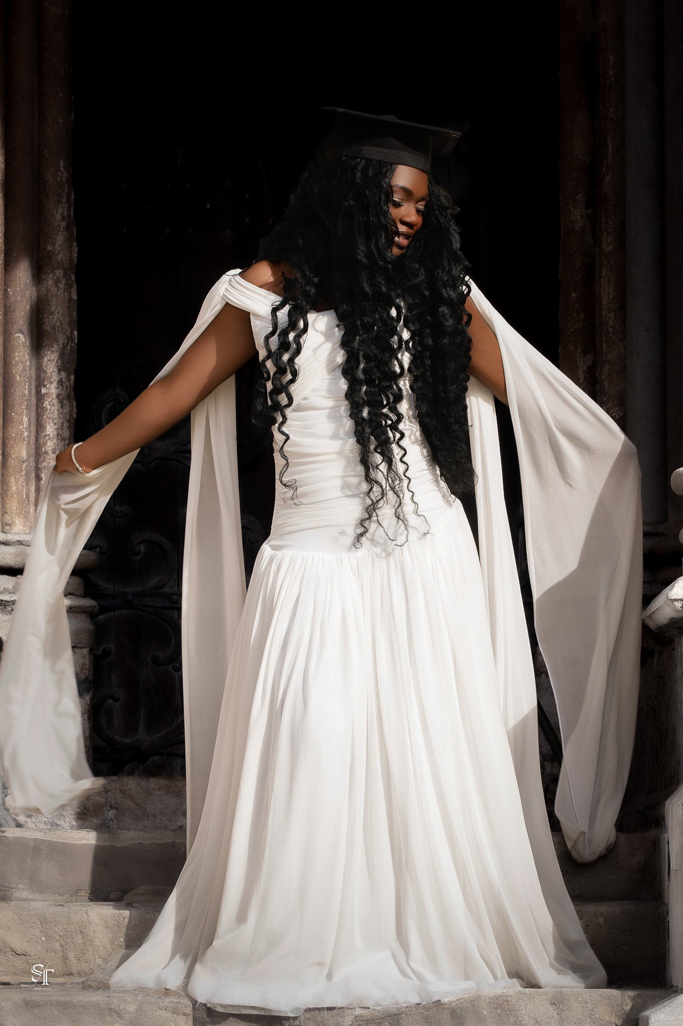 Woman in a white wedding dress and black graduation cap standing on stairs in front of a stone wall.
