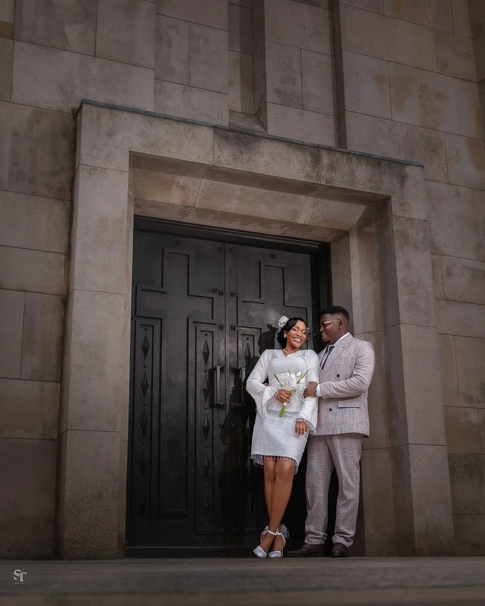 A smiling couple dressed in wedding attire standing in front of a large black door in a stone building, holding a bouquet of white flowers.