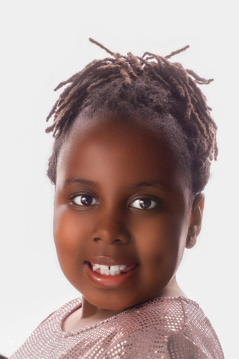 Portrait of a young girl with brown skin, brown eyes, and short dreadlocks, smiling at the camera, wearing a pink mesh top against a white background.