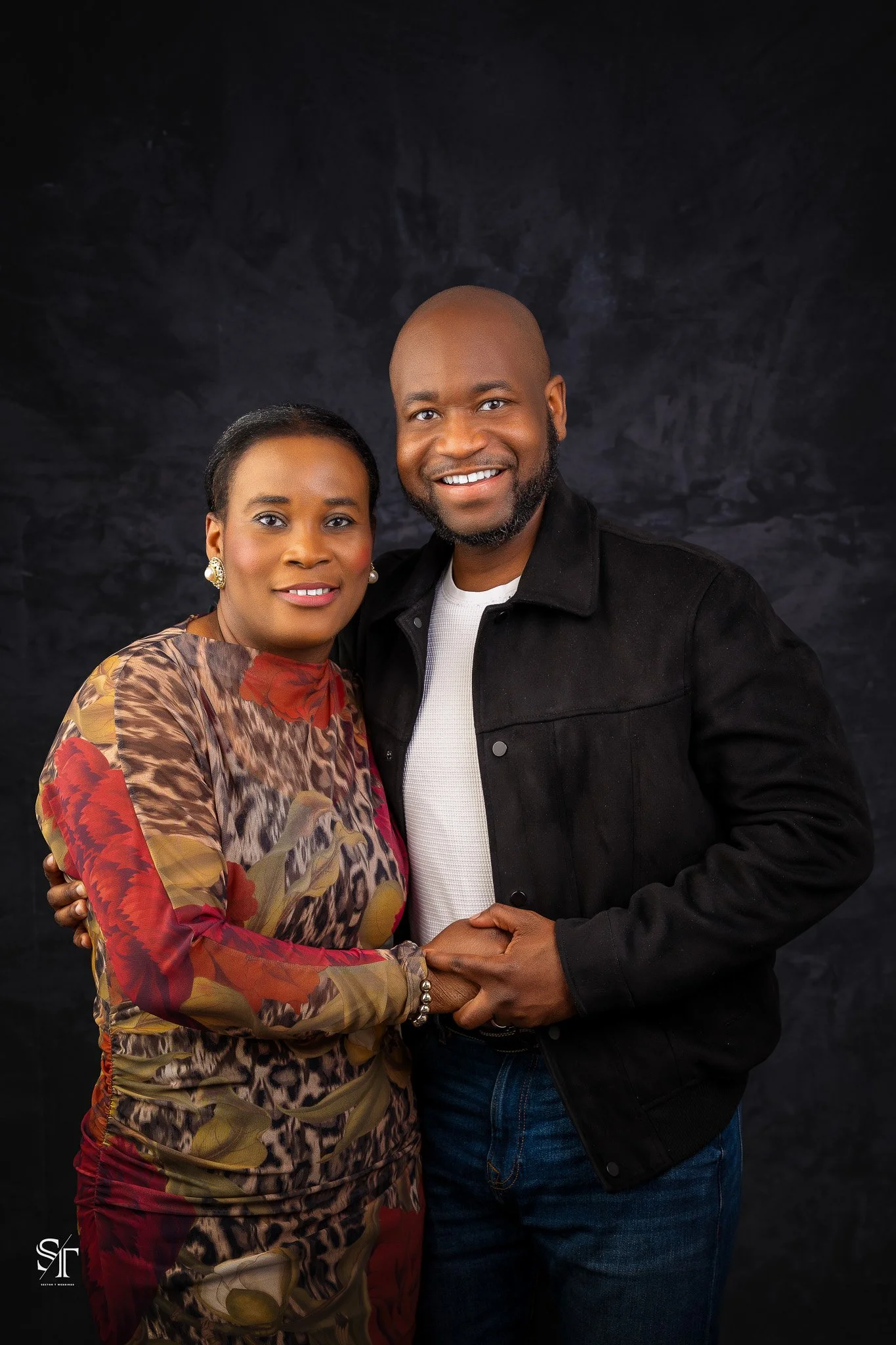 A smiling Black couple holding hands and looking at the camera, standing close against a dark backdrop.