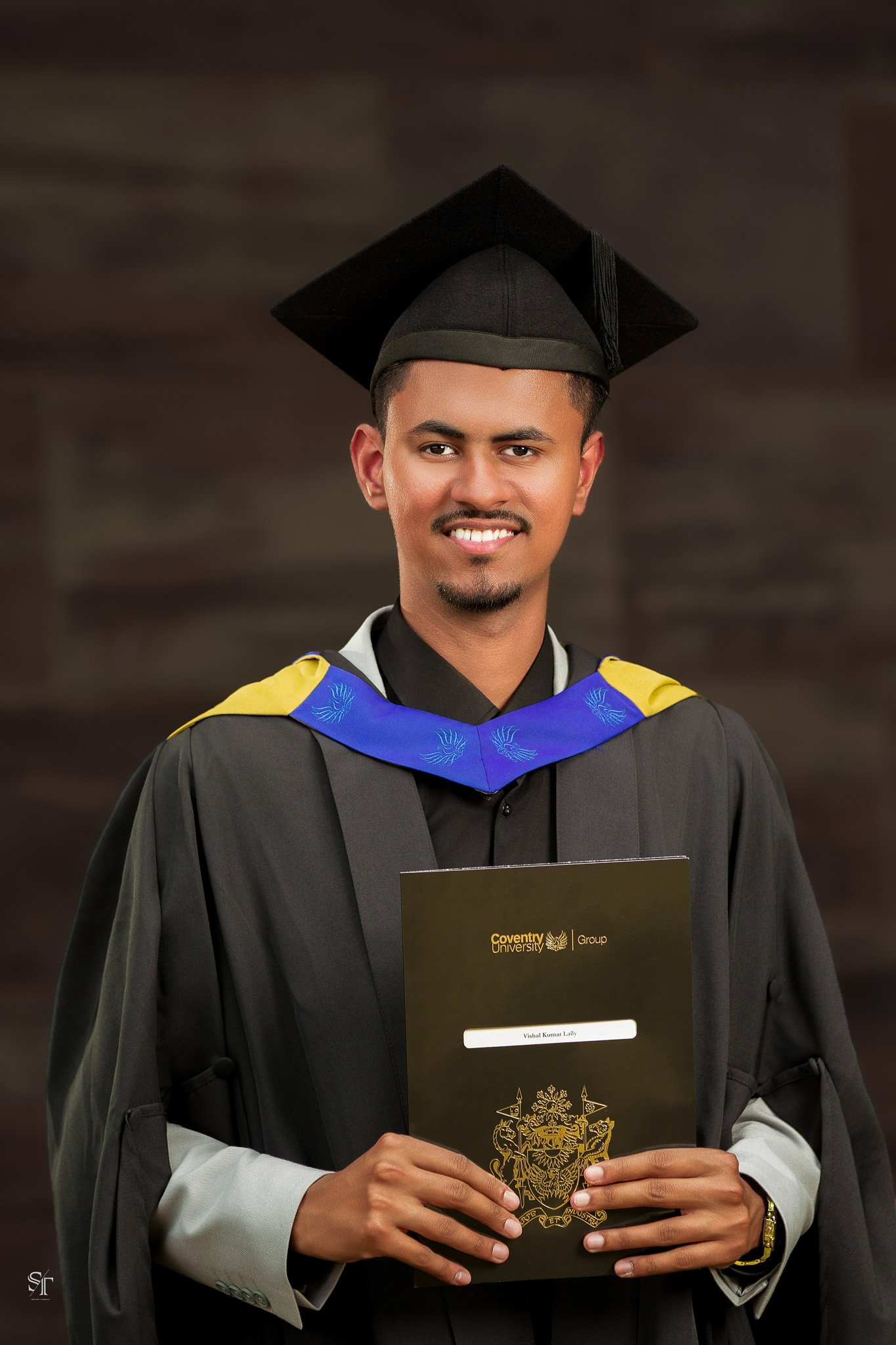 Young man in graduation gown and cap holding a certificate, smiling, with a dark background.