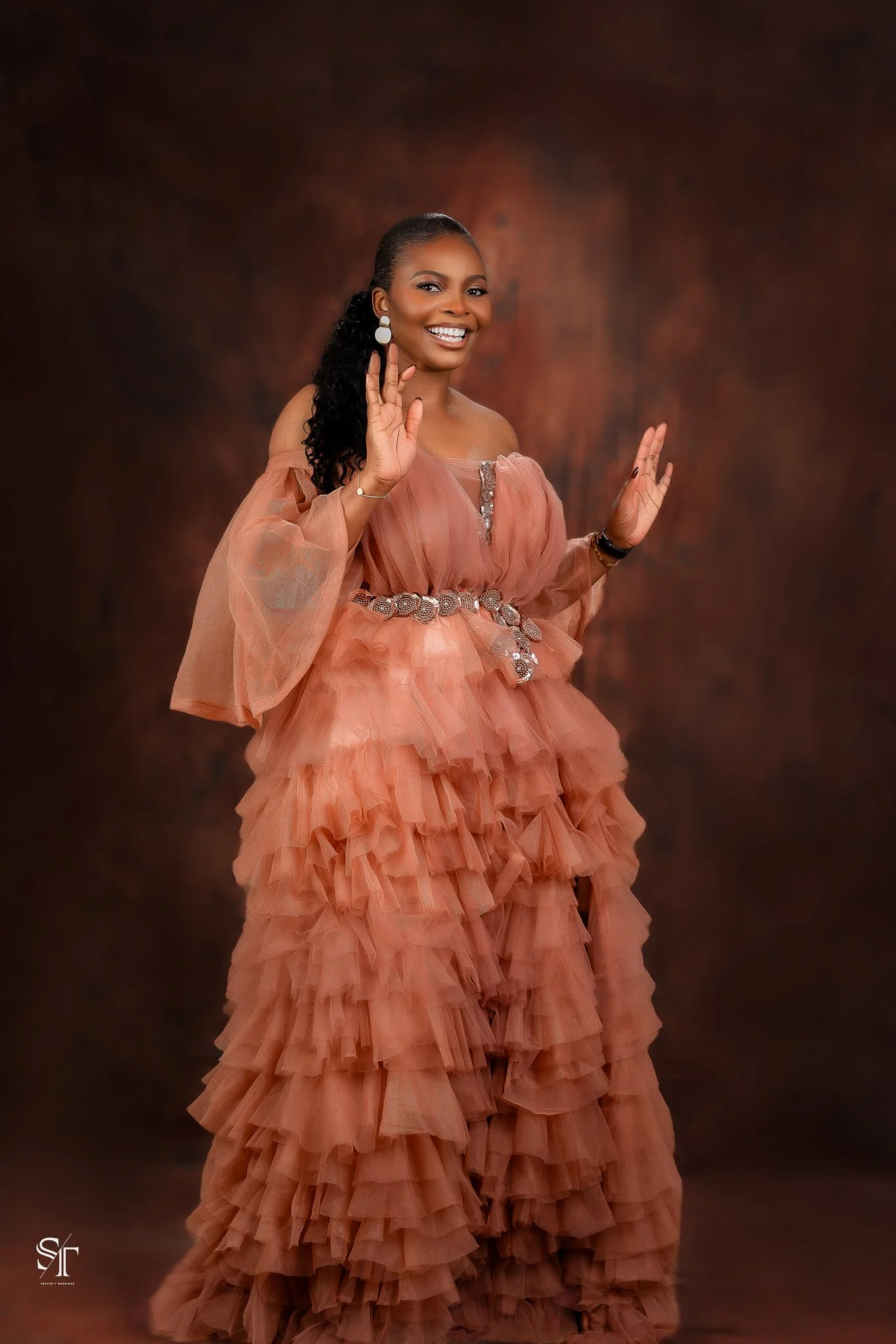 A woman dressed in a peach-colored layered tulle dress with puffed sleeves, smiling and holding her hands up in a friendly gesture against a dark brown background.