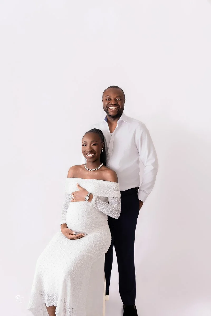 A smiling pregnant woman in a white off-shoulder dress and a pearl necklace, sitting and holding her belly, with a smiling man in a white shirt and black pants standing beside her against a plain white background.