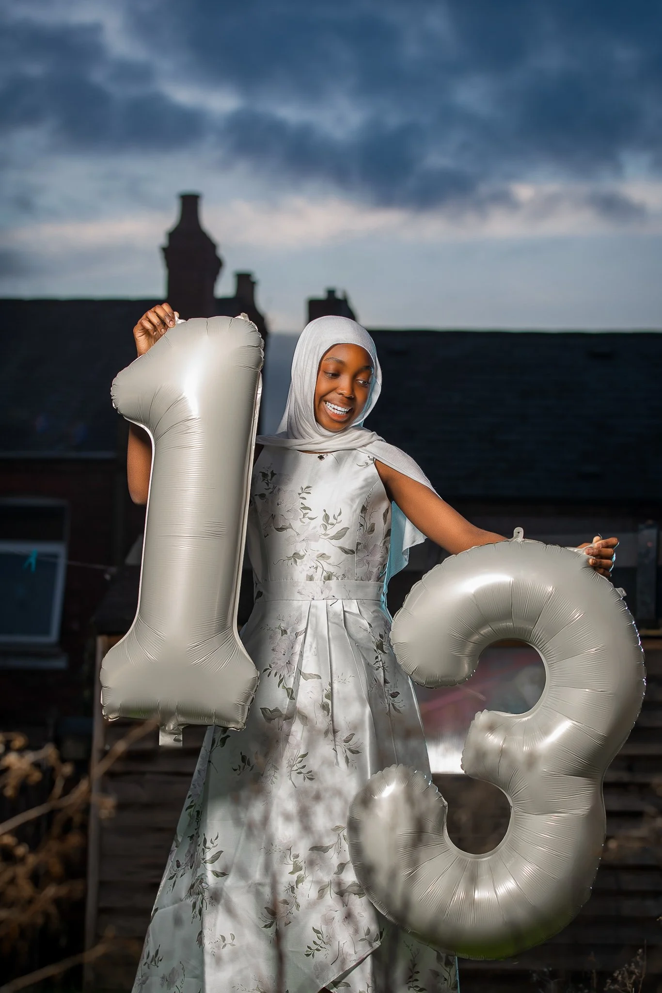 A woman wearing a floral dress and hijab holding silver balloons shaped as the numbers 1, 3, and 2, celebrating a 132nd birthday or milestone outdoors during twilight.
