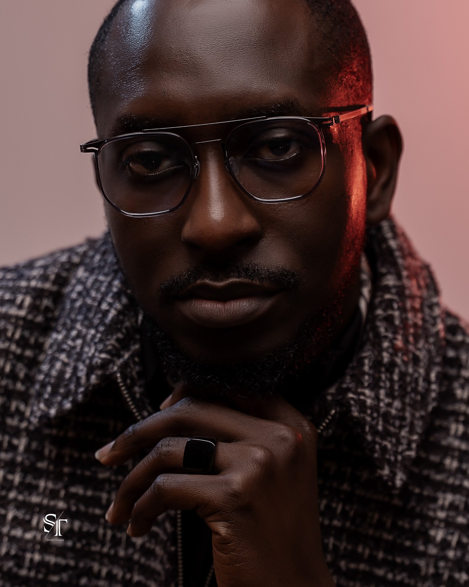 Close-up portrait of a person with dark skin wearing glasses, a patterned jacket, and a ring, with a pink background and soft lighting.