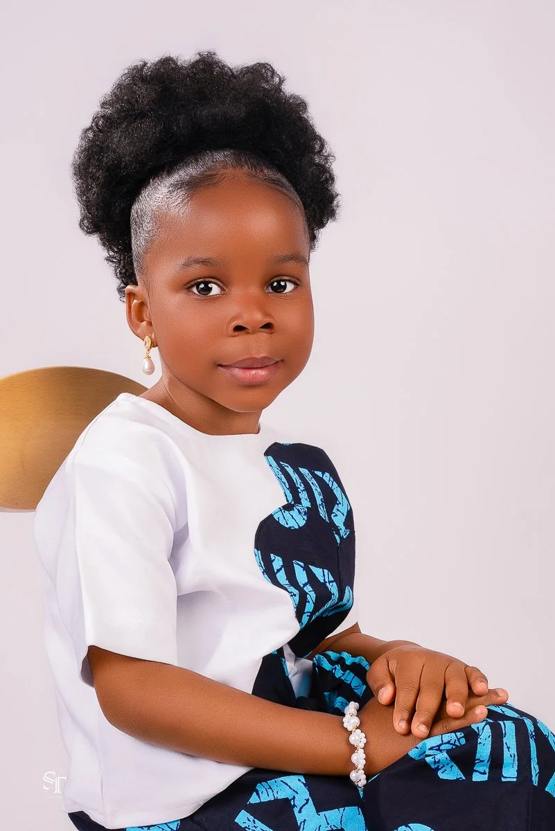 A young girl with dark curly hair styled in a puff, wearing a white shirt with a large blue and black design, gold earrings with pearl accents, and a white beaded bracelet, sitting on a wooden chair against a plain white background.