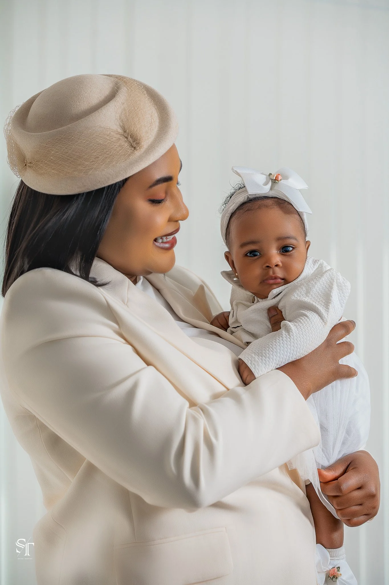 A woman in a cream-colored blazer and hat holding a young girl in a white dress with a bow on her head.