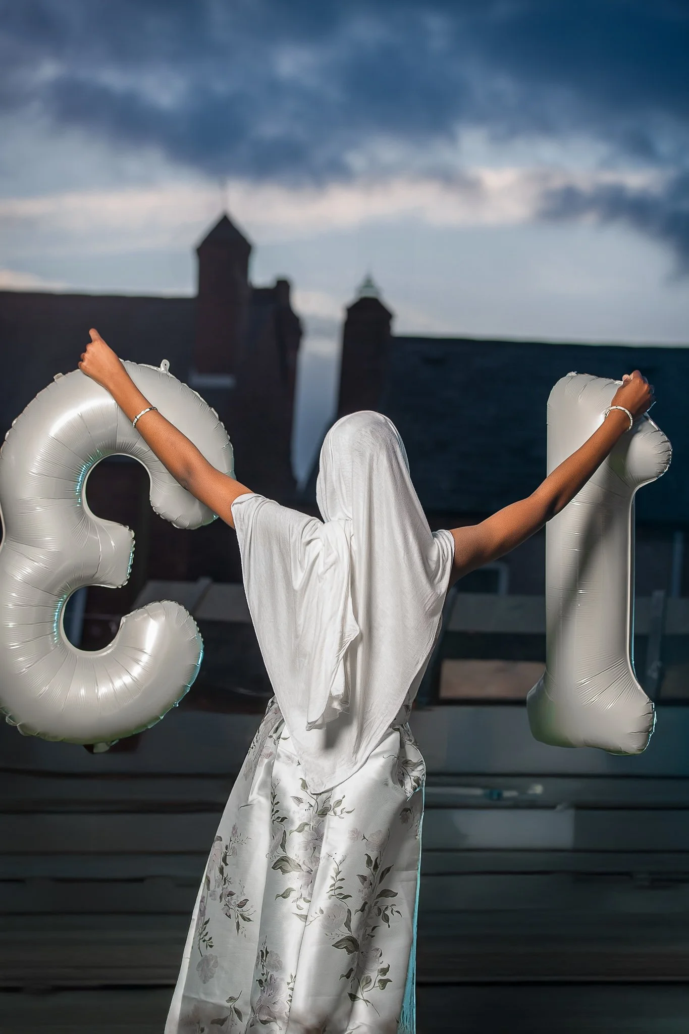 A person dressed in white with a cloth covering their head, holding large silver balloon letters 'C' and 'L' in an outdoor setting during twilight.