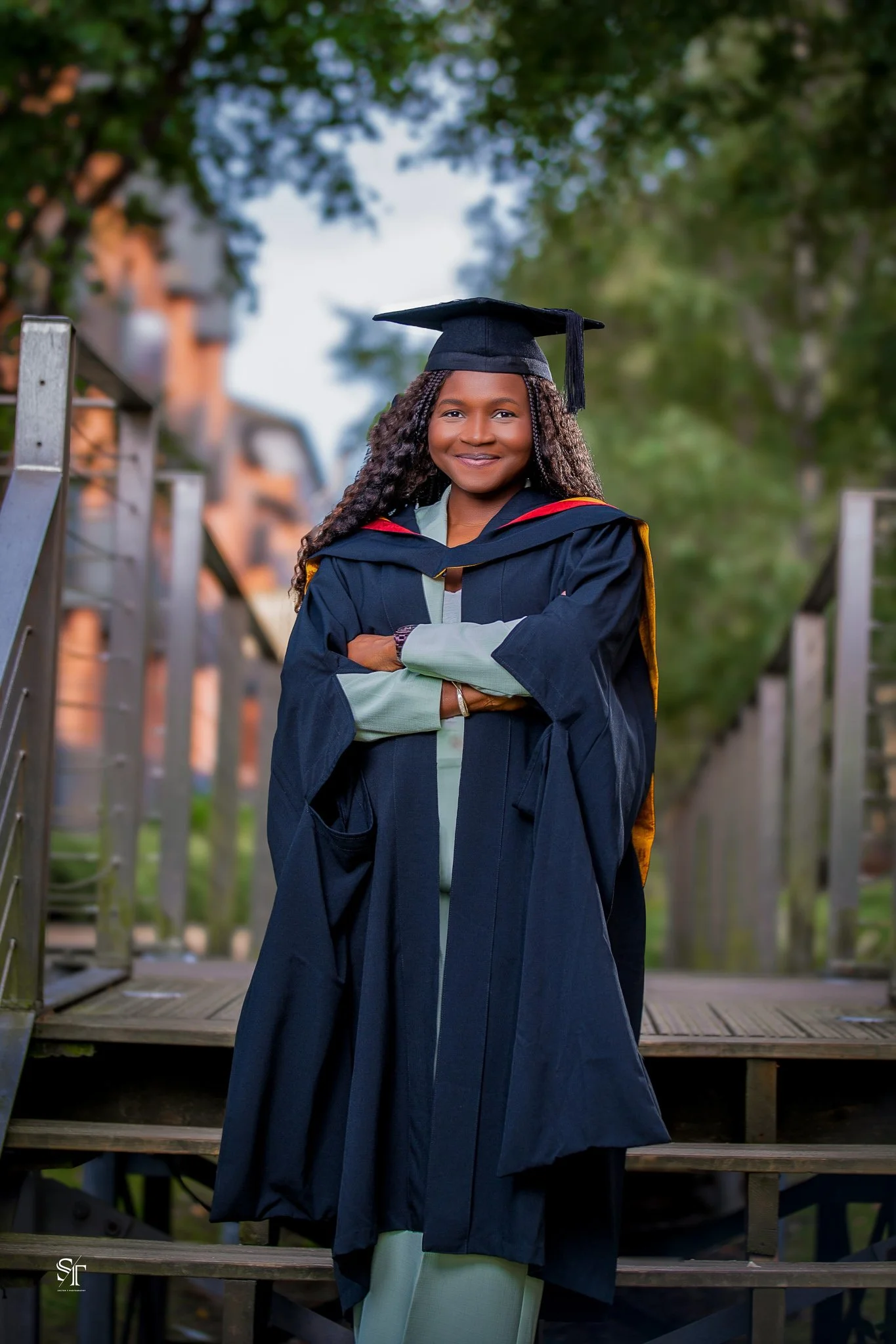 A young woman in graduation cap and gown standing outdoors on wooden steps with trees in the background, smiling with arms crossed.
