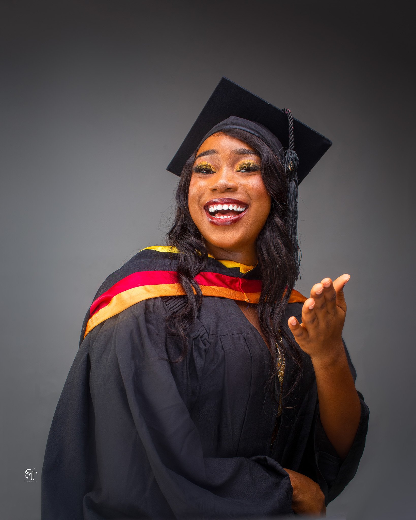 A young woman in a graduation cap and gown smiling and looking at the camera against a dark gray background.
