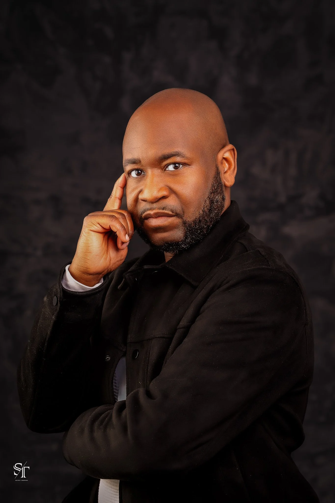 A bald African American man with a beard, wearing a black jacket, poses with his hand touching his temple against a dark background.