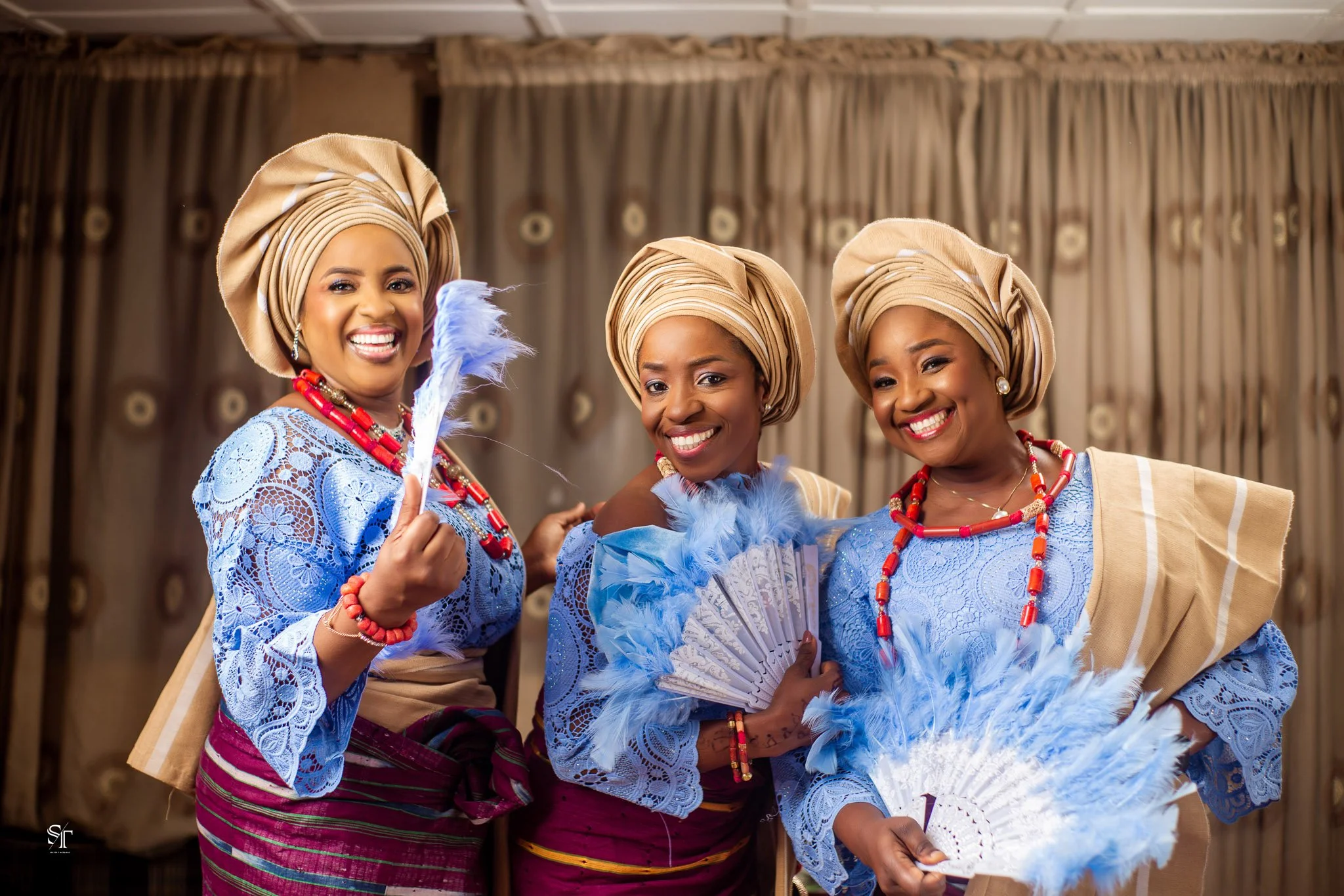 Three women dressed in traditional Nigerian attire smiling and holding fans, standing in front of a curtain backdrop.